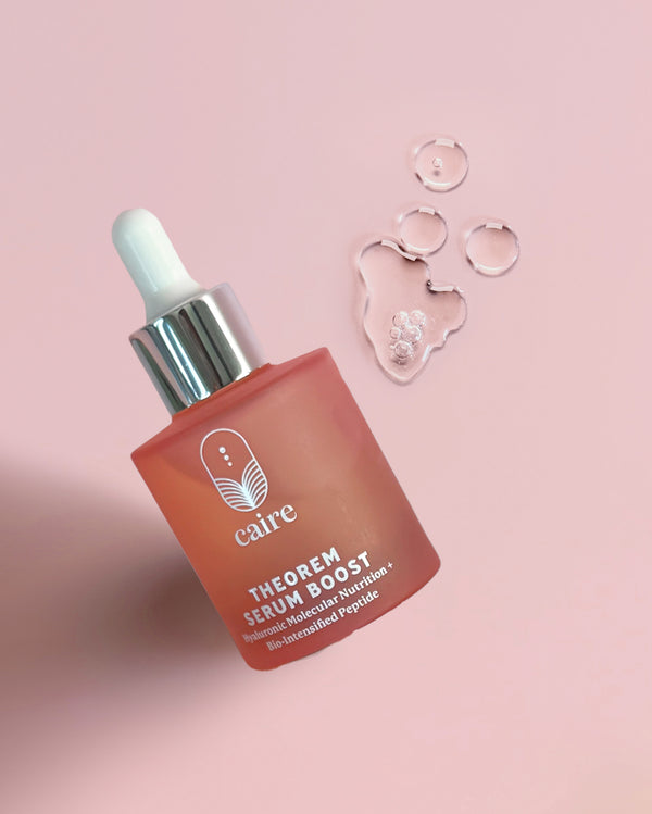 A bottle of Cairé Theorem Serum Boost with a dropper cap, set on a soft pink background with clear liquid droplets and a heart-shaped gel piece beside it.