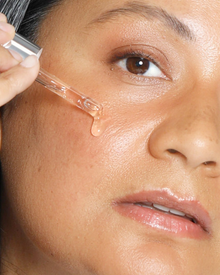 Close-up of a person applying a clear skincare serum or dropper liquid to their cheek, with a single drop hanging from the glass pipette.