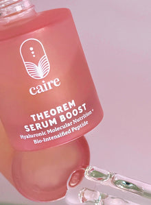 A close-up of a pink bottle labeled 'caire THEOREM SERUM BOOST' with a glass pipette resting near the base, against a soft pink background.
